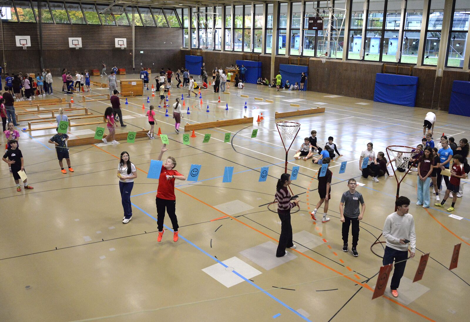 Blick in die große Sporthalle: Viele Schülerinnen und Schüler verteilen sich auf Stationen mit Hütchen, Reifen und Bänken.