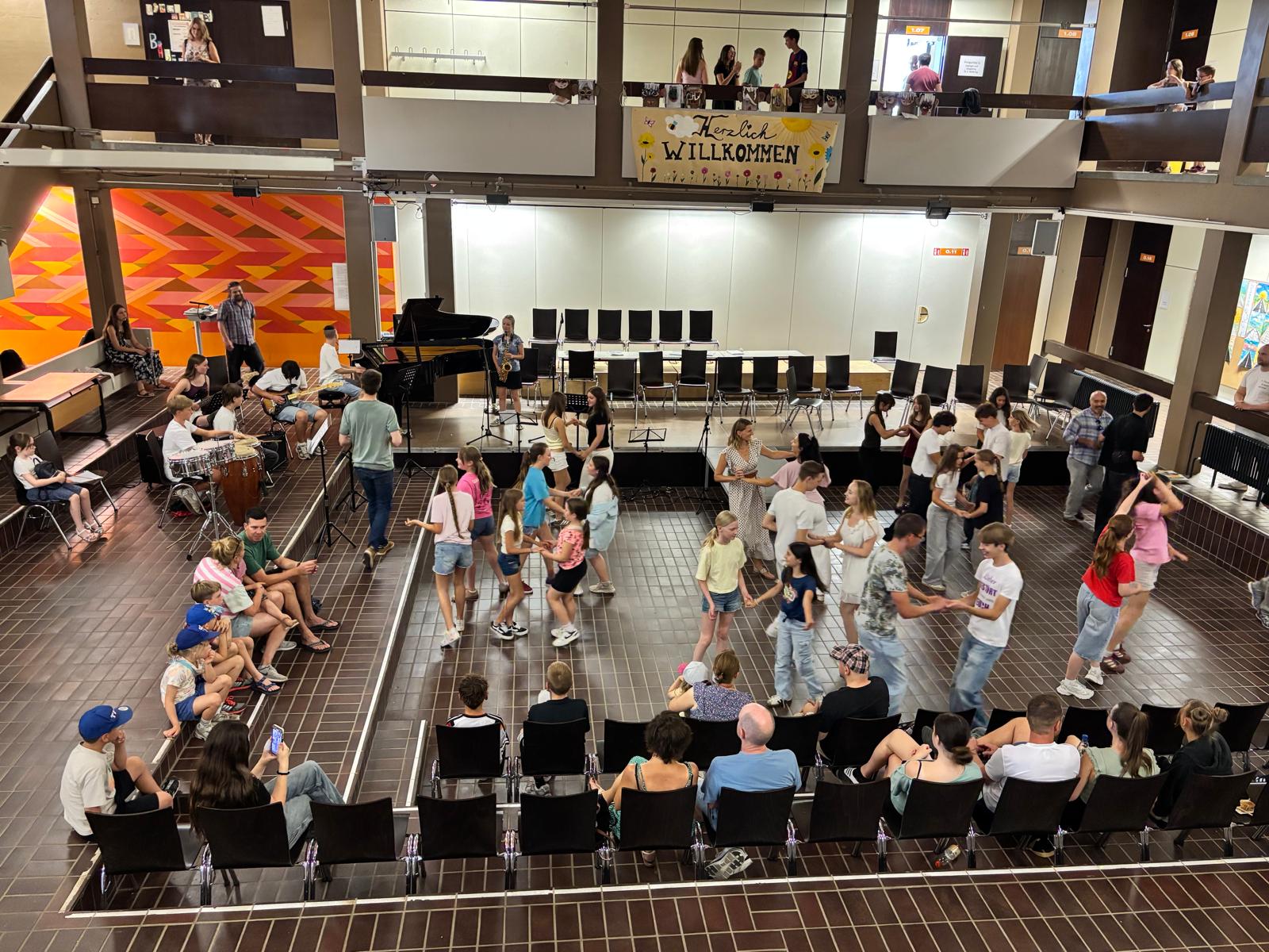 Blick von der Galerie in die Aula: Schülerinnen und Schüler tanzen paarweise, eine Schulband mit Flügel, Saxofon, Gitarre und Trommeln spielt dazu. Gäste sitzen auf Stühlen und Stufen, oben hängt ein Banner mit 'Herzlich Willkommen'.