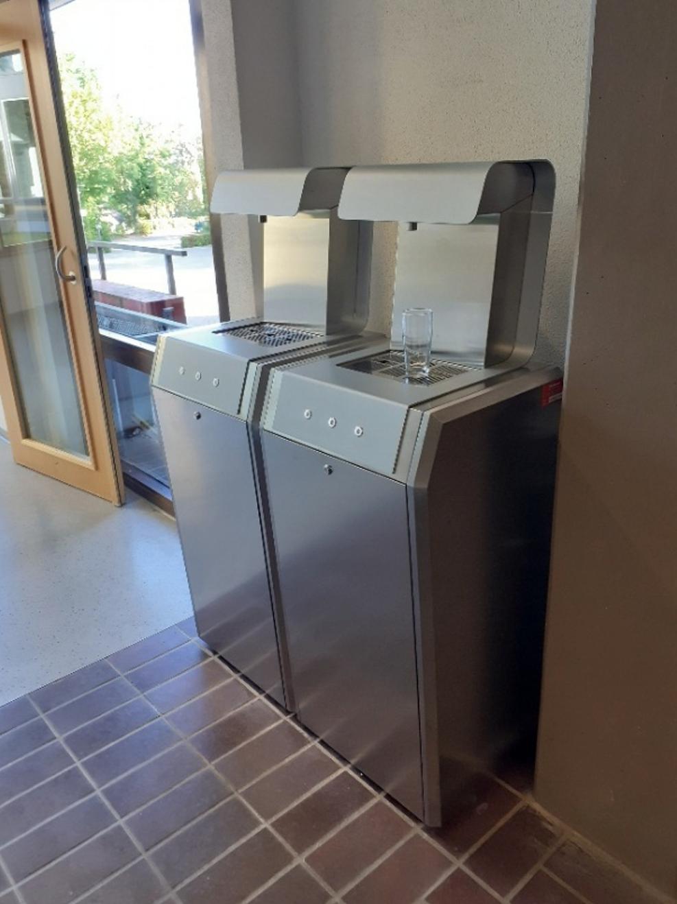 Zwei nebeneinanderstehende Trinkwasserspender aus Edelstahl mit Tasten und Abtropfgitter, auf einem steht ein gefülltes Wasserglas. Daneben eine offene Glastür mit Blick nach draußen.
